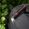 Olight Perun 2 Mini ブラックとレッドのL型ヘッドライトをクリップでグレーのバッグに装着し、屋外の草花に光を当てるクローズアップシーン